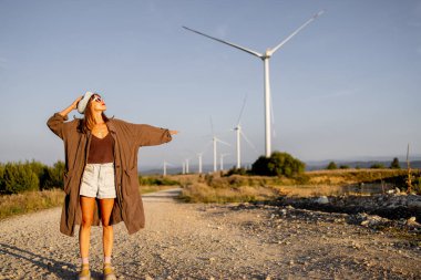 Arka planda rüzgar türbinleriyle kırsal bir yolda duran bir kadın esintiyi hissetmek için kollarını açıyor. Temiz enerjinin ve çevre dostu yaşamın geleceğini kucaklıyor.