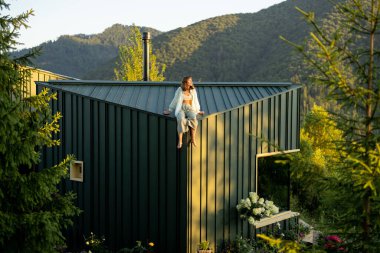 A woman enjoys a tranquil morning on a mountain cabin rooftop, surrounded by lush nature and stunning views, embodying rest, relaxation, and a deep connection with the wilderness. clipart