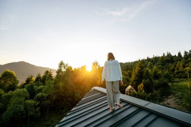 Güneş dağların üzerinden yükselirken, doğanın huzur dolu yalnızlığını ve güzelliğini kucaklayan bir kadın çatıda yürür. Mükemmel bir sakinlik ve yansıma anı.