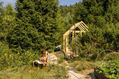 Female carpenter building a wooden greenhouse in her backyard on a sunny summer day. Concept of DIY projects and sustainable living. Vide view on mountains clipart