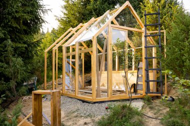 Uzun ağaçlarla çevrili verimli bir arka bahçede yapım aşamasındaki ahşap bir sera. Sahne, sürdürülebilir yaşam formlarını ön plana çıkaran, çevre dostu bir ortamda DIY projesinin ilk aşamalarını yakalıyor.