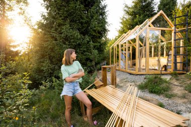 Female carpenter standing next to a partially built wooden greenhouse in her backyard, wearing casual clothes and work gloves, showcasing a DIY project in a nature-filled environment clipart