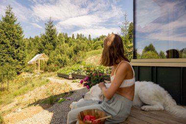 A woman sits on the porch, savoring a snack while her dog rests peacefully by her side. The lush greenery and clear skies create a tranquil atmosphere clipart