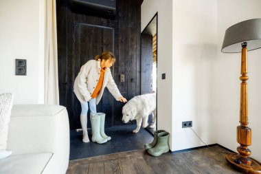 Kapalı kabinde rahat bir kadın köpeğini kapı eşiğine yerleştiriyor, botları yerde, sıcak ve sade bir kırsal atmosfer sergiliyor.