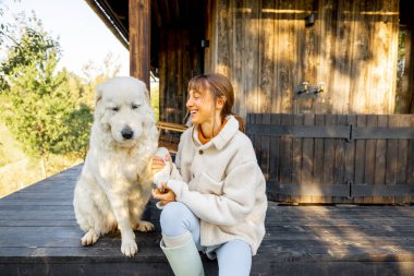 Ahşap bir verandada oturan bir kadın köpeğinin patisini tutuyor, ikisi de açık hava ortamının tadını çıkarıyor. Kırsal bir ortamda insan ve evcil hayvan arasındaki bağı yansıtan neşeli ve sıcak bir an.