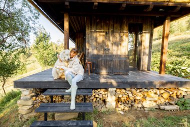 Verandada oturan bir kadın büyük beyaz köpeğine sarılıyor, dışarıda sıcak bir anı paylaşıyor. Ahşap veranda ve doğal ortam, arkadaşlığı ve doğayı yansıtan kırsal bir cazibe ekler.