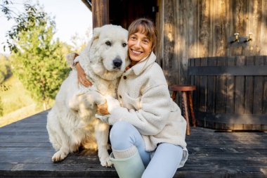 Verandada oturan bir kadın büyük beyaz köpeğine sarılıyor, dışarıda sıcak bir anı paylaşıyor. Ahşap veranda ve doğal ortam, arkadaşlığı ve doğayı yansıtan kırsal bir cazibe ekler.