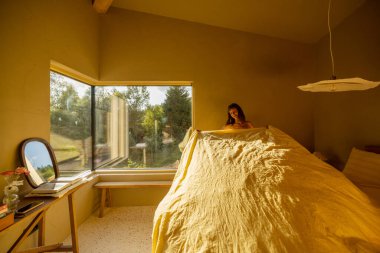 Ilık, sarı bir yatak odasında yumuşak sarı bir yatak örtüsüyle yatağı yapan bir kadın. Büyük pencere, odayı doğal ışıkla dolduran yemyeşil bir manzara sunuyor.