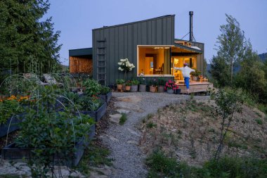 Günbatımında modern kabin, yumuşak, sıcak iç aydınlatma ile rahat yaşam alanını aydınlatıyor. Çiçekli güzel bahçe, dağın etrafındaki huzuru tamamlıyor.