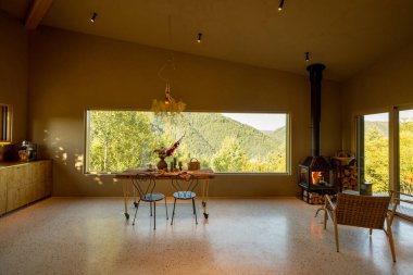 Geniş bir pencerenin önünde süslü ahşap kahvaltı masası manzarası ve yemyeşil bir dağ manzarası sergiliyor.