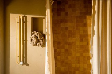 Yumuşak bej fayansların ve doğal kumaş perdelerin bulunduğu güneşli bir duş alanı sıcak, samimi bir his yaratıyor. Havlu kancaları ve desenli duvarlar biraz kırsal konfor katıyor.