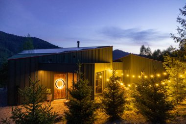 Alacakaranlıkta sıcak sicim ışıklarıyla aydınlatılan, alacakaranlık gökyüzüne karşı çam ağaçlarıyla çevrili, rahat bir dağ kulübesinin dış görünüşü.