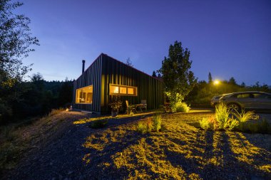 A modern cabin illuminated at twilight with a soft glow from interior lights. Surrounded by nature, a parked car and garden landscaping add to the serene and cozy atmosphere clipart