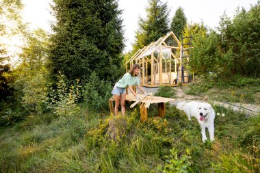 Bir tezgahın yanında duran kadın marangoz ve arka bahçesinde kısmen inşa edilmiş ahşap bir seranın önünde duran büyük beyaz köpeği. Doğa ile çevrili bir DIY projesiyle meşgul.
