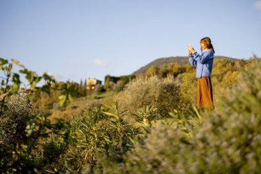 Yeşillik ve sıcak güneş ışığı altında yuvarlanan tepelerle çevrili Toskana kırsalının manzaralı bir fotoğrafını çeken bir kadın. Seyahat, doğa ve kırsal cazibe temaları için mükemmel.