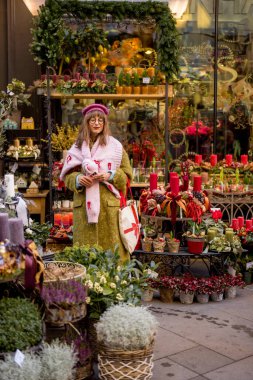 Kış kıyafetleri içinde şık bir kadın bir butik dükkanının önünde canlı tatil süslemeleriyle çevrili duruyor. Çelenkler, çiçekler ve mumlar sıcak, şenlikli bir ortam yaratır.