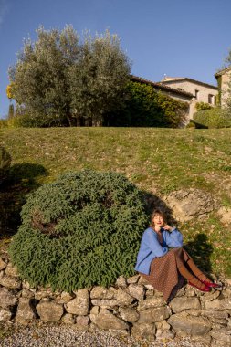 Güneşli bir günde yemyeşil ve zeytin ağaçlarıyla çevrili kırsal taştan bir duvarda oturan bir kadın kırsal kesimin huzurlu güzelliğinin tadını çıkarıyor.