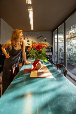 Minimalist elbiseli bir kadın kırsal bir yemek masasını çiçeklerle süslüyor. Büyük cam pencereler yumuşak doğal ışık getirir, sıcak ve huzurlu atmosferi güçlendirir.