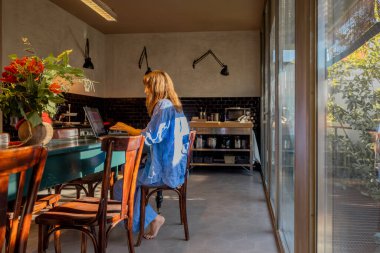 Rahat mavi elbiseli çıplak ayaklı bir kadın modern bir mutfakta laptopuyla kırsal bir yemek masasında çalışıyor. Güneş ışığı büyük cam pencerelerden geçer, rahat ve ilham verici bir çalışma alanı yaratır. 