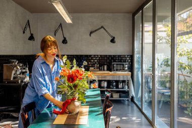 Rahat mavi gömlekli bir kadın şık bir mutfakta kırsal bir yemek masasını çiçeklerle süslüyor. Büyük cam pencerelerden gelen doğal ışık sıcak ve davetkar atmosferi güçlendirir.