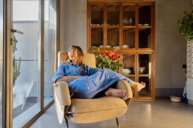 Mavi keten elbiseli bir kadın, güneşli bir pencerenin yanındaki eski bir koltukta dinleniyor. Modern iç mekan, ahşap dolap ve taze çiçekler sıcak, rahat ve ilham verici bir yaşam ortamı yaratır.