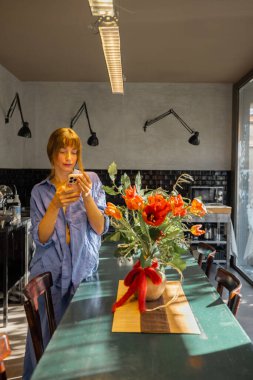 Rahat mavi elbiseli bir kadın kırsal bir yemek masasında duruyor, modern bir mutfakta akıllı telefonunu kontrol ediyor. Büyük cam pencereler sıcak doğal ışık getirir, rahat ve zarif bir atmosfer yaratır.