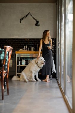 Çıplak ayaklı bir kadın modern bir mutfakta duruyor, elini nazikçe büyük beyaz köpeğinin üzerinde dinlendirirken kahvesini yudumluyor. Cam pencerelerden gelen doğal ışık huzurlu ve rahat atmosferi güçlendirir.