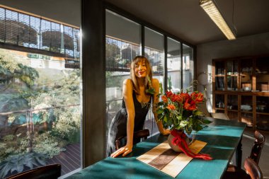 Neşeli bir kadın kırsal bir yemek masasına dayanıyor ve modern bir mutfakta çiçek aranjmanı yapıyor. Büyük cam pencereler boşluğu sıcak doğal ışıkla doldurur, sıcak ve davetkar bir ortam yaratır.