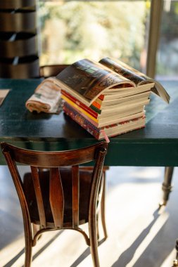 Renkli kitaplarla dolu rahat bir köşe ve ahşap bir masanın üzerinde açık bir dergi. Güneş ışığı pencereden süzülerek incelenmek için mükemmel sıcak ve ilham verici bir atmosfer yaratır.