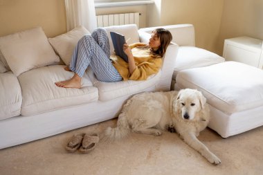 Sıcak bir kazak ve çizgili pijama pantolonlu rahat bir kadın yumuşak bir halıya uzanmış kitap okurken pofuduk beyaz köpeğine sarılıyor. Evde huzurlu bir rahatlık ve arkadaşlık anı.