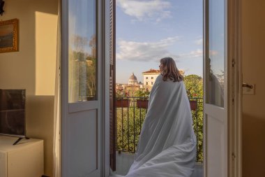 Beyaz bir battaniyeye sarılmış bir kadın rahat bir balkonda duruyor Vatikan 'daki St. Peters Bazilikası' nın büyüleyici sabah manzarasına bakıyor.