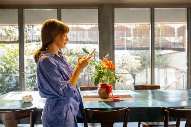 Rahat mavi elbiseli çıplak ayaklı bir kadın, akıllı telefonunu kullanarak kırsal bir yemek masasının yanında duruyor. Büyük cam pencereler şık alanı doğal ışıkla doldurur, huzurlu ve rahat bir atmosfer yaratır. 