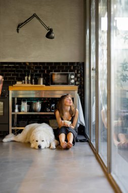 Çıplak ayaklı bir kadın modern bir mutfağın zemininde oturmuş büyük beyaz köpeğinin yanında kahvesini yudumluyor. Cam pencerelerden gelen doğal ışık uzayın huzurlu ve rahatlatıcı atmosferini arttırır.