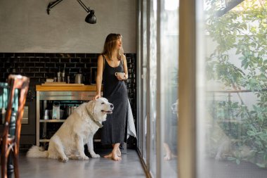 Çıplak ayaklı bir kadın modern bir mutfakta duruyor, elini nazikçe büyük beyaz köpeğinin üzerinde dinlendirirken kahvesini yudumluyor. Cam pencerelerden gelen doğal ışık huzurlu ve rahat atmosferi güçlendirir.