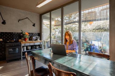 Rahat mavi kıyafetli bir kadın modern bir mutfakta laptopuyla kırsal bir yemek masasında çalışıyor. Geniş cam pencereler boşluğu doğal ışıkla doldurarak sakin ve üretken bir atmosfer yaratıyor.