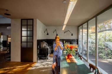 Rahat mavi elbiseli bir kadın kırsal bir yemek masasında duruyor, modern bir mutfakta akıllı telefonunu kontrol ediyor. Büyük cam pencereler sıcak doğal ışık getirir, rahat ve zarif bir atmosfer yaratır.
