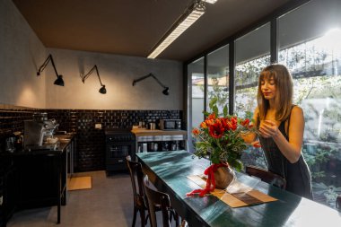 Bir kadın, koyu fayanslı ve büyük cam pencereli şık bir mutfakta kırsal bir yemek masasını çiçeklerle süslüyor. Yumuşak doğal ışık hoş ve zarif atmosferi güçlendirir.