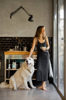 Çıplak ayaklı bir kadın modern bir mutfakta duruyor, elini nazikçe büyük beyaz köpeğinin üzerinde dinlendirirken kahvesini yudumluyor. Cam pencerelerden gelen doğal ışık huzurlu ve rahat atmosferi güçlendirir.