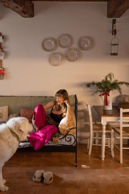 Rahatlatıcı, sade, ahşap kirişler ve klasik dekorla döşenmiş bir ev. Parlak pantolonlu bir kadın demir bir yatakta dinlenir, yumuşak güneş ışığıyla yıkanır.. 