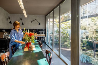 Rahat mavi gömlekli bir kadın şık bir mutfakta kırsal bir yemek masasını çiçeklerle süslüyor. Büyük cam pencerelerden gelen doğal ışık sıcak ve davetkar atmosferi güçlendirir.