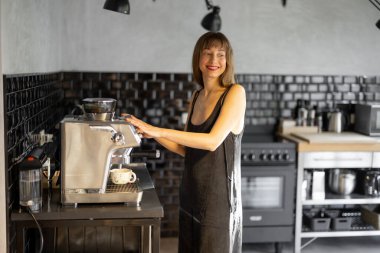 Minimalist elbiseli neşeli bir kadın şık bir mutfakta profesyonel bir espresso makinesiyle kahve hazırlıyor. Koyu karolar ve sıcak doğal ışık rahat ve zarif bir atmosfer yaratır.