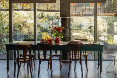 Ahşap sandalyeli güzel bir yemek masası, şenlikli çiçek aranjmanları, ve geniş pencerelere bakan yemyeşil kitaplar. Doğal ışık ve rahat atmosferin karışımı...