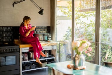 Pembe elbiseli bir kadın mutfak tezgahında oturup telefonuyla sessiz bir sabahın tadını çıkarıyor. Güneş ışığı cam kapılardan geçer, sıcak ve huzurlu bir atmosfer yaratır.