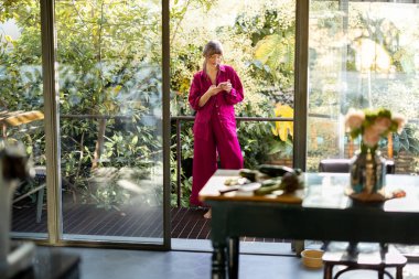 Pijamalı bir kadın terasta çıplak ayakla duruyor. Etrafı yeşilliklerle çevrili, telefonunu çeviriyor. Doğal ortamda sakin bir an, güne duyarlı bir başlangıcı simgeliyor.