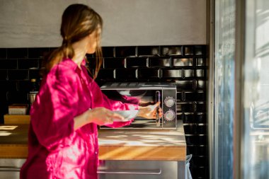 Pembe elbiseli bir kadın kahvaltı hazırlar, tabağı modern bir tezgah fırınına koyar. Güneş ışığıyla yıkanan siyah fayanslı mutfak sıcak ve minimalist bir sabah atmosferi yaratır.