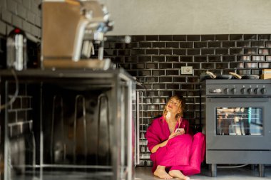 Pembe elbiseli bir kadın mutfak zemininde çıplak ayakla oturur, telefonu tutarken yukarı bakar. Siyah fayanslı iç mekan ve sıcak aydınlatma modern yaşamın rahat ve yansıtıcı atmosferini yaratır.