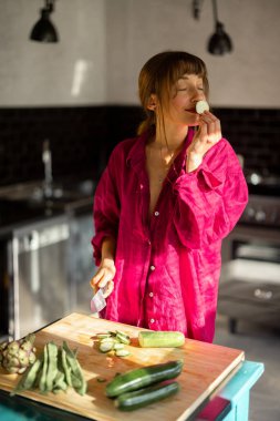 Rahat bir ev ortamında bir kadın, sıcak doğal ışıkla yıkanmış, ahşap bir masada taze sebzeler hazırlıyor. Yavaş yaşamın, sağlığın ve düşünceli günlük ritüellerin bir temsili.