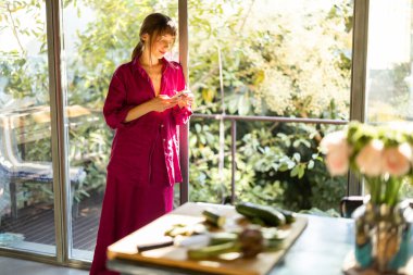 Rahat pijamalı bir kadın, güneşli bir mutfakta sessiz bir sabah geçirir ve telefonunu kontrol eder. Sıcak doğal ışık ve yeşillik huzurlu ve yavaş yaşayan bir atmosfer yaratır.