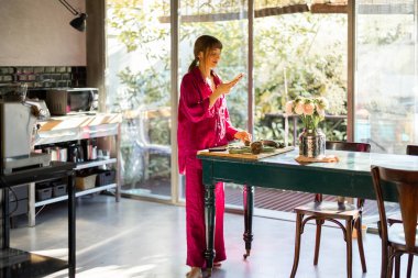 Yumuşak pijamalı bir kadın, güneşli mutfağında huzurlu bir sabah geçirir ve taze sebzeler hazırlarken telefonundan tarifini kontrol eder. Sahne yavaş yaşam ve dikkatli rutinleri somutlaştırıyor.