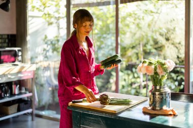 Rahat bir ev ortamında bir kadın, sıcak doğal ışıkla yıkanmış, ahşap bir masada taze sebzeler hazırlıyor. Yavaş yaşamın, sağlığın ve düşünceli günlük ritüellerin bir temsili.